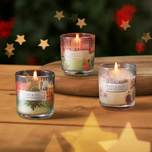 Three lit Heart & Home mini candles in illustrated Christmas glass holders on a wooden board with soft star bokeh in the background.