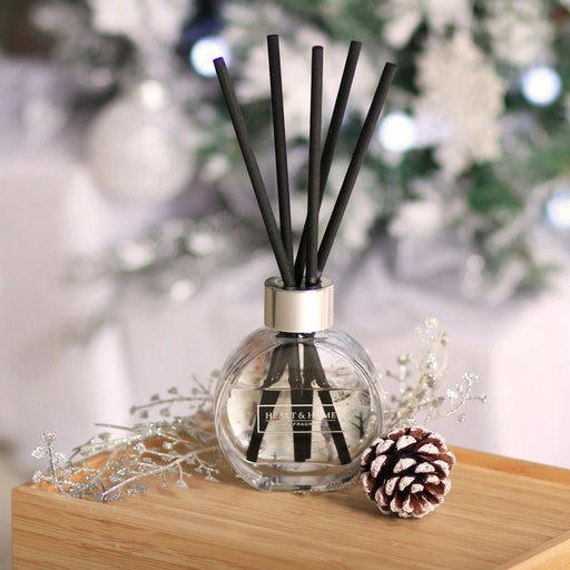 Round bauble-style reed diffuser with chunky black reeds on a wooden shelf, frosty greenery and winter lights softly blurred in the background.
