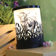 Highland cow silhouette electric wax melt burner lit at an angle on a wooden surface with small flowers nearby.