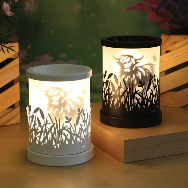 Two electric wax melt burners lit in a styled setting, one black and one white, both with a Highland cow silhouette design.