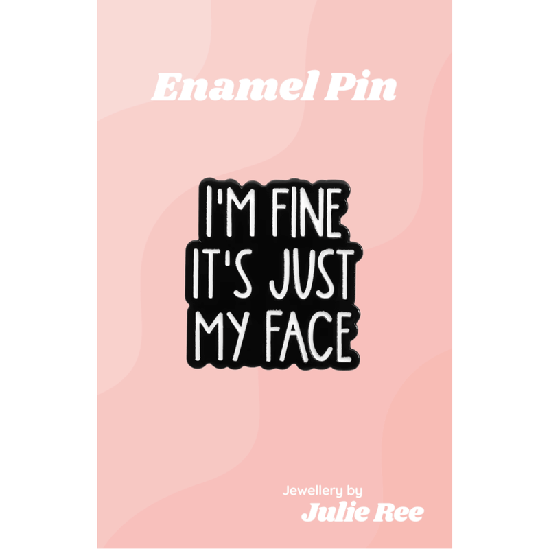 Black enamel pin with white text reading I’m fine it’s just my face, displayed on a pink backing card.