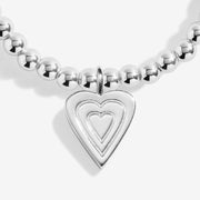 Close‑up of polished layered‑heart pendant hanging from petite round silver beads on stretchy bracelet.
