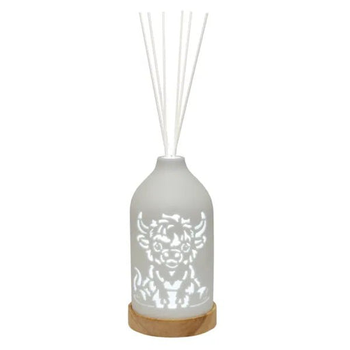 White LED ceramic reed diffuser with a Highland cow cut-out design on a wooden base with white reed sticks.