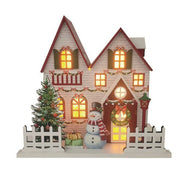 White wooden Christmas house with red roof, lit windows, wreath, tree and snowman, 24 x 22cm, on white background.