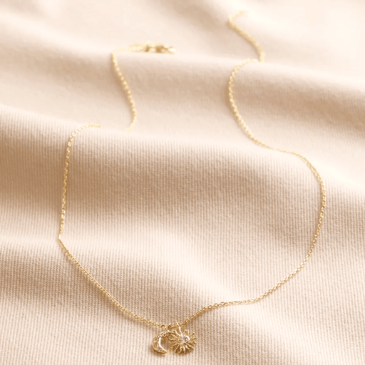 Gold sun and moon charm necklace laid on fabric, featuring a sunburst charm and crescent moon with cubic zirconia crystals.