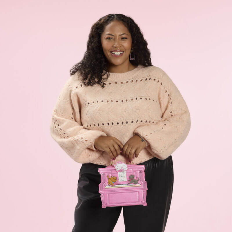 Model holding the Loungefly Disney The Aristocats figural piano crossbody bag by the top handle, shaped like a pink piano.