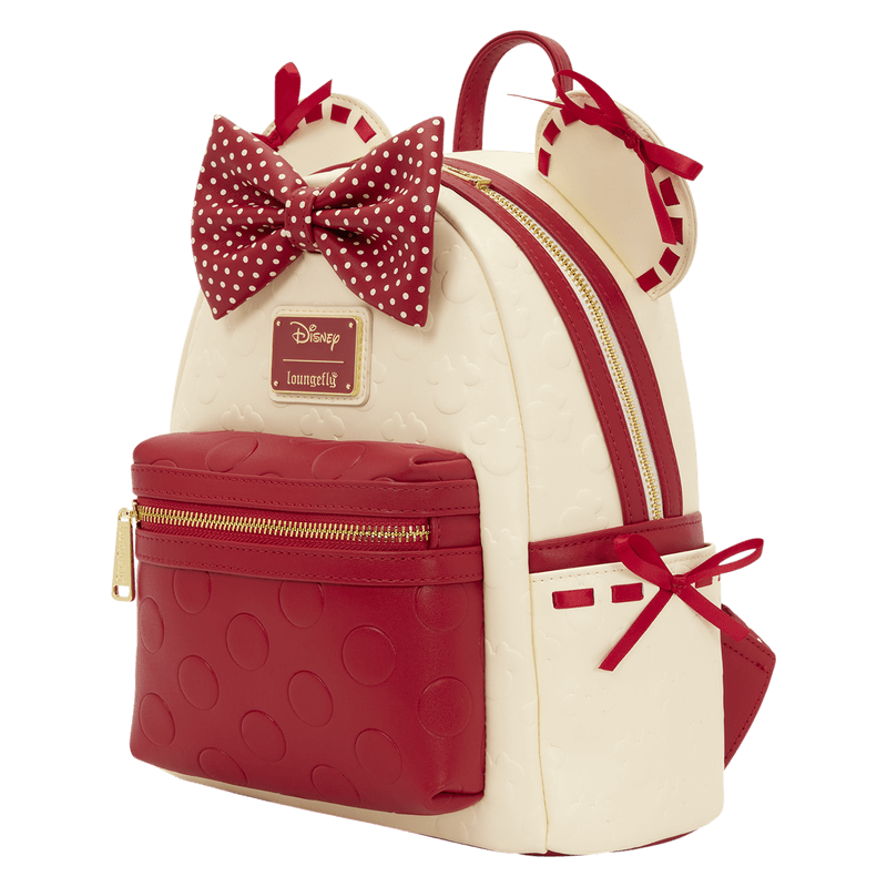 Angled view of the Loungefly Disney Minnie Mouse mini backpack showing the red polka dot bow, gold zip, cream body and red front pocket.
