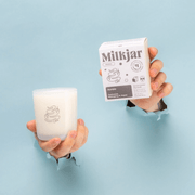 Hands holding Milkjar Aurora candle and box against a blue background.