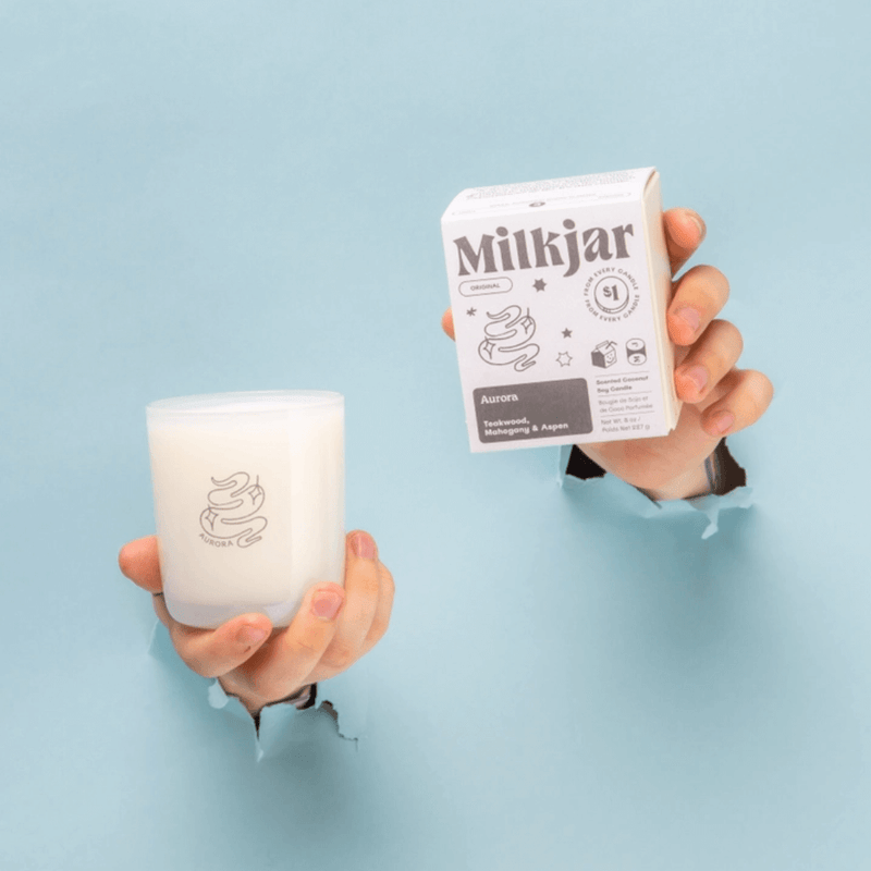 Hands holding Milkjar Aurora candle and box against a blue background.