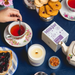 Overhead view of Milkjar Blueberry Tea candle and box beside a cup of tea, biscuits and toast with jam on a blue tablecloth.