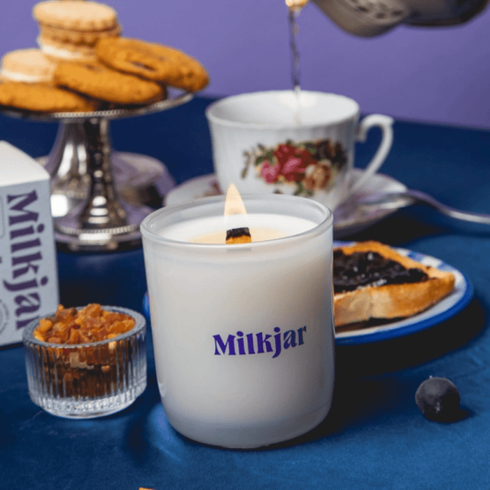Close-up of lit Milkjar Blueberry Tea coconut soy candle with crackling wooden wick on a tea table with teacup and treats.