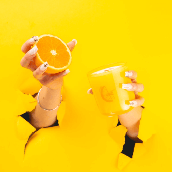 Hands holding an orange and Milkjar Citrus candle through torn yellow paper, bright yellow backdrop.