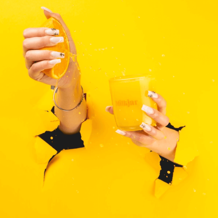 Hands squeezing an orange next to a Milkjar Citrus candle against a bright yellow background.