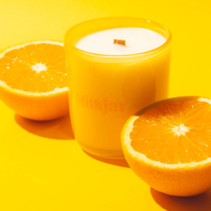 Milkjar Citrus candle in yellow glass surrounded by orange halves on a yellow background.
