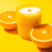Milkjar Citrus candle in yellow glass surrounded by orange halves on a yellow background.