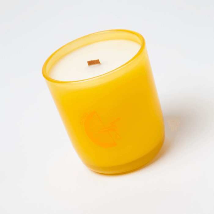 Angled view of Milkjar Citrus candle in yellow glass with wooden wick on white background.