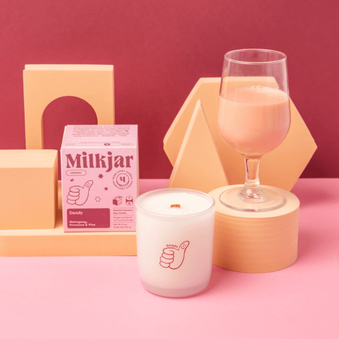 Milkjar Dandy candle and box styled on a pink set with a glass drink and geometric props in the background.