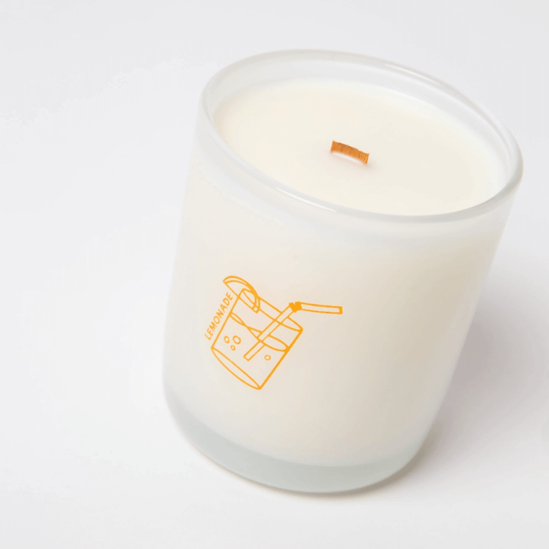 Close-up angled view of Milkjar Lemonade candle in frosted glass with wooden wick and lemonade icon on a white background.