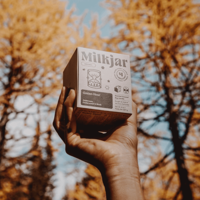 Hand holding Milkjar Golden Hour candle box outdoors with autumn trees in the background.