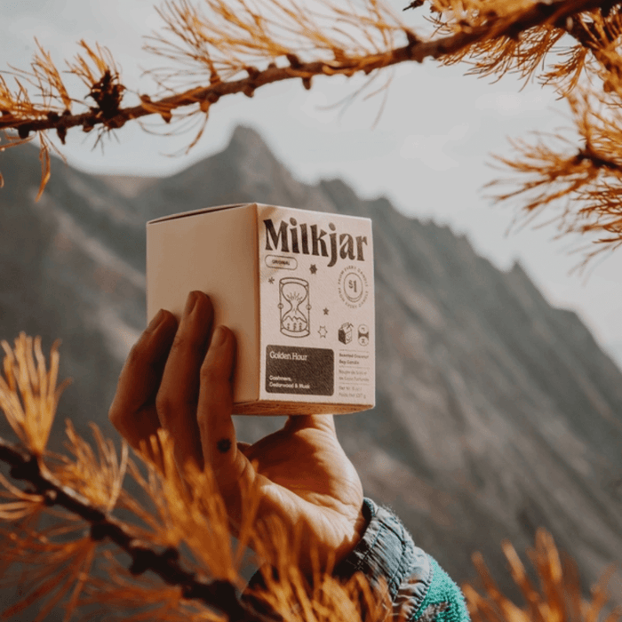 Hand holding Milkjar Golden Hour candle box outdoors with mountain scenery behind.