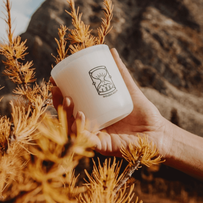 Hand holding Milkjar Golden Hour candle outdoors among golden leaves and rocky landscape.
