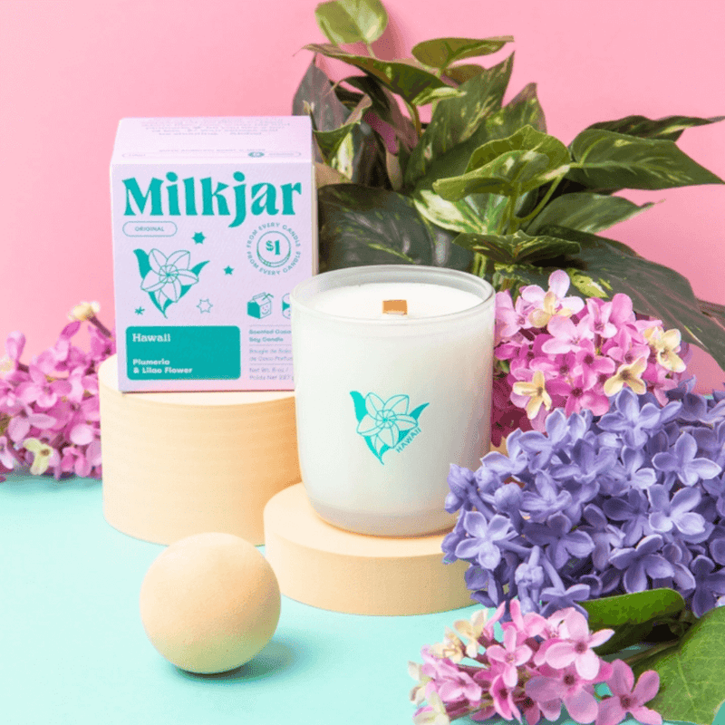 Milkjar Hawaii candle and box styled with flowers, green leaves and pastel props on a pink background.