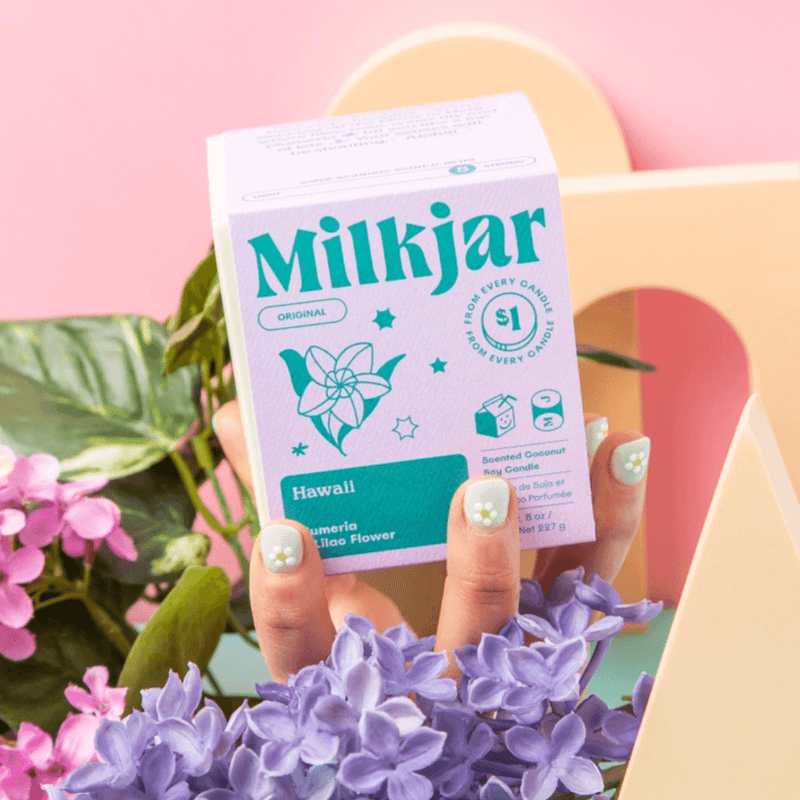 Close-up of hands holding the Milkjar Hawaii candle box with flowers in the foreground on a pink background.