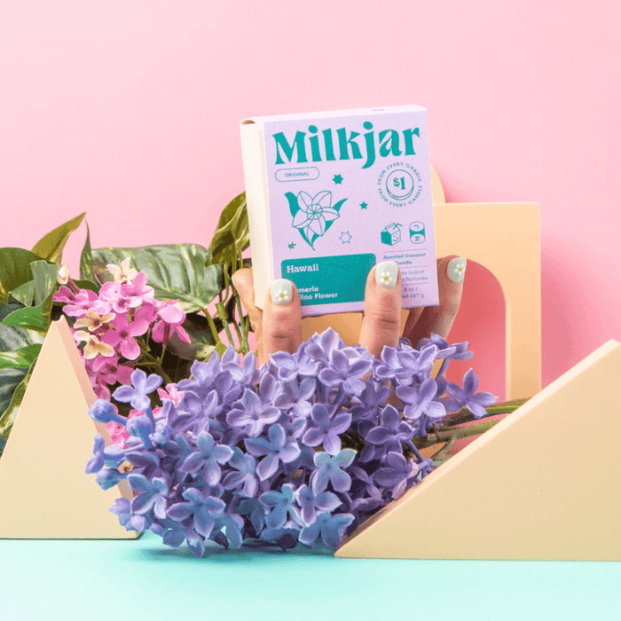 Hand holding the Milkjar Hawaii frosted glass candle among flowers and pastel props on a pink background.
