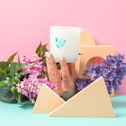 Hand holding the Milkjar Hawaii candle on a pastel set with flowers and geometric props.