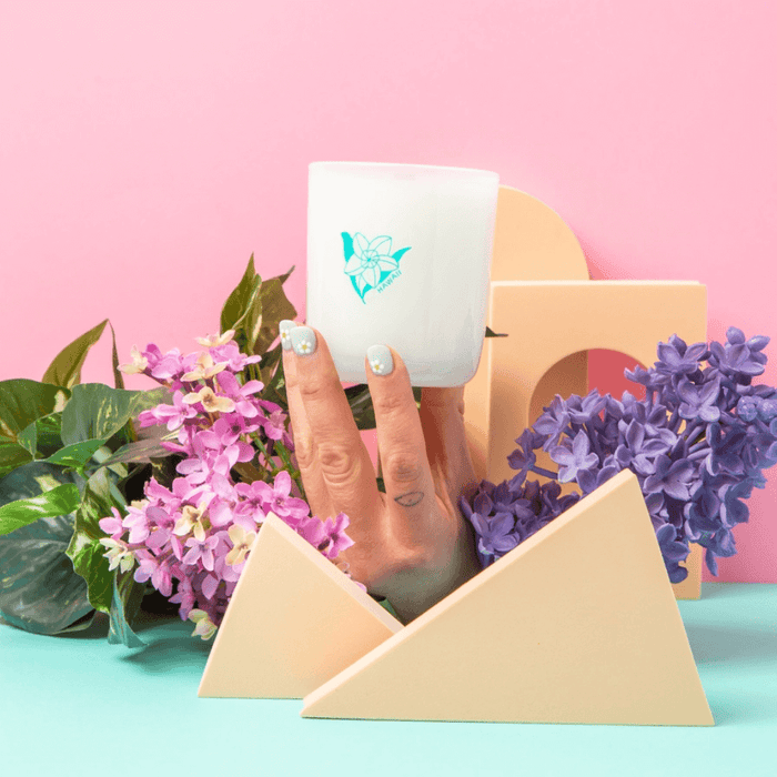 Hand holding the Milkjar Hawaii candle on a pastel set with flowers and geometric props.