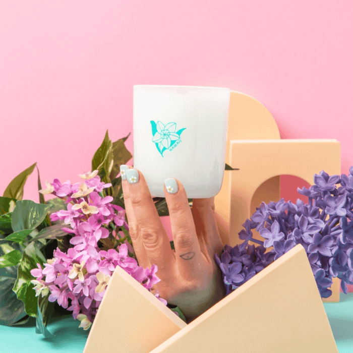 Hand holding the Milkjar Hawaii frosted glass candle among flowers and pastel props on a pink background.