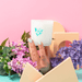 Hand holding the Milkjar Hawaii frosted glass candle among flowers and pastel props on a pink background.