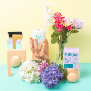 Wide styled scene of Milkjar Hawaii candle and box with flowers, pastel props and a hand holding the candle.