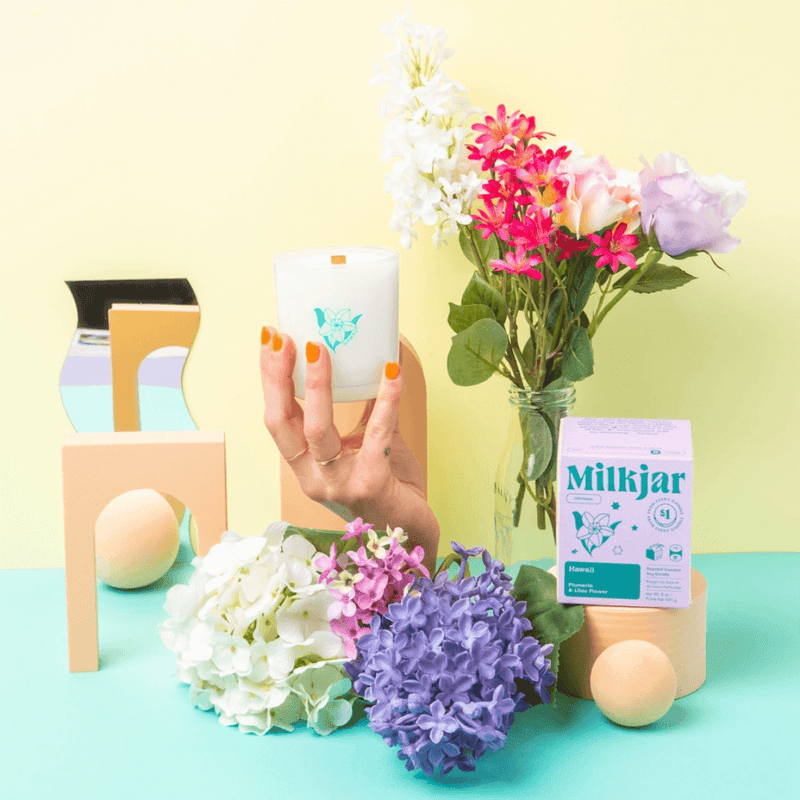 Wide styled scene of Milkjar Hawaii candle and box with flowers, pastel props and a hand holding the candle.