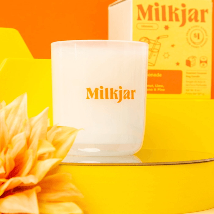 Close-up of Milkjar Lemonade candle with Milkjar branding on a bright yellow set with the candle box in the background.