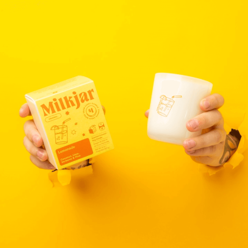 Hands holding Milkjar Lemonade candle and box through torn yellow paper on a bright yellow background.