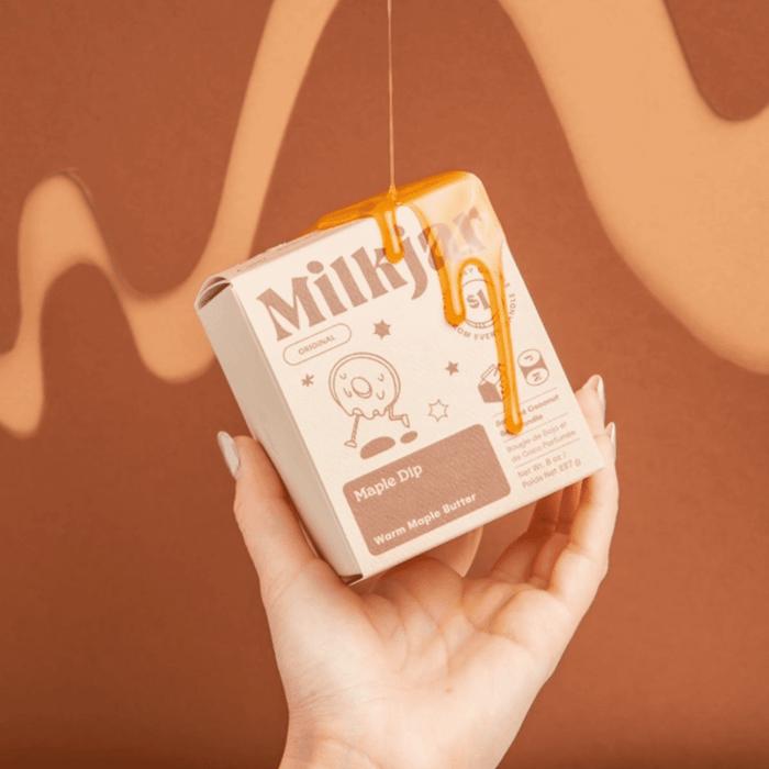 Hand holding Milkjar Maple Dip candle box with maple syrup dripping over the packaging on a warm brown backdrop.