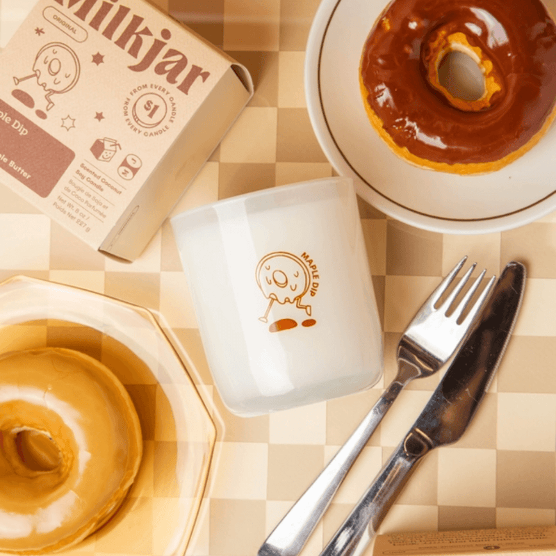 Overhead flatlay of Milkjar Maple Dip candle with doughnuts, cutlery and the Maple Dip box on a beige checkered surface.