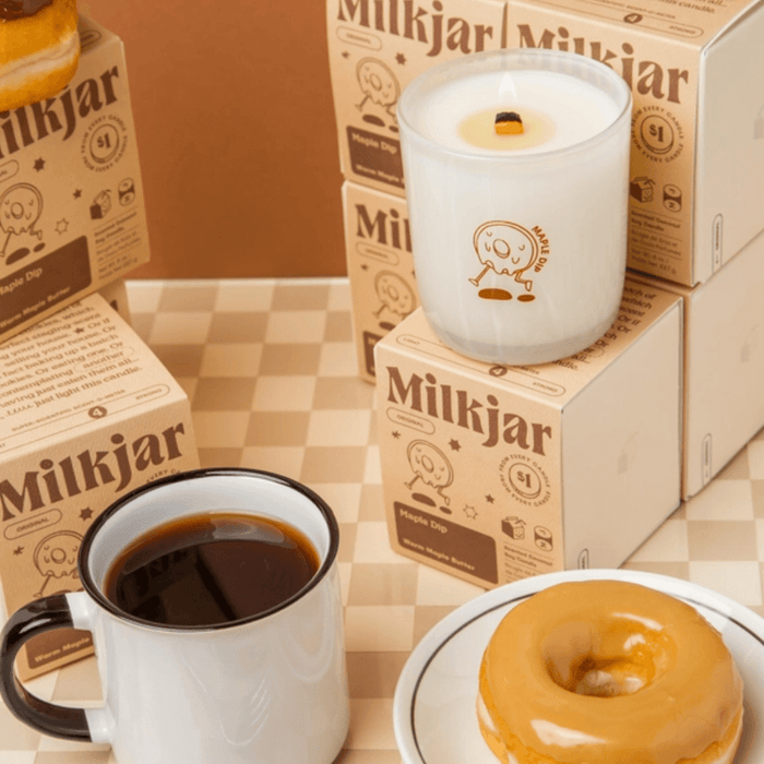 Lit Milkjar Maple Dip candle on stacked boxes beside a mug of coffee and glazed doughnut on a checkered table.