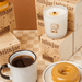 Lit Milkjar Maple Dip candle on stacked boxes beside a mug of coffee and glazed doughnut on a checkered table.