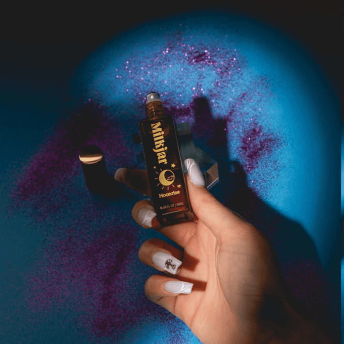 Hand holding Milkjar Moonrise Neroli & Plum 20ml perfume roller against blue and purple glitter background