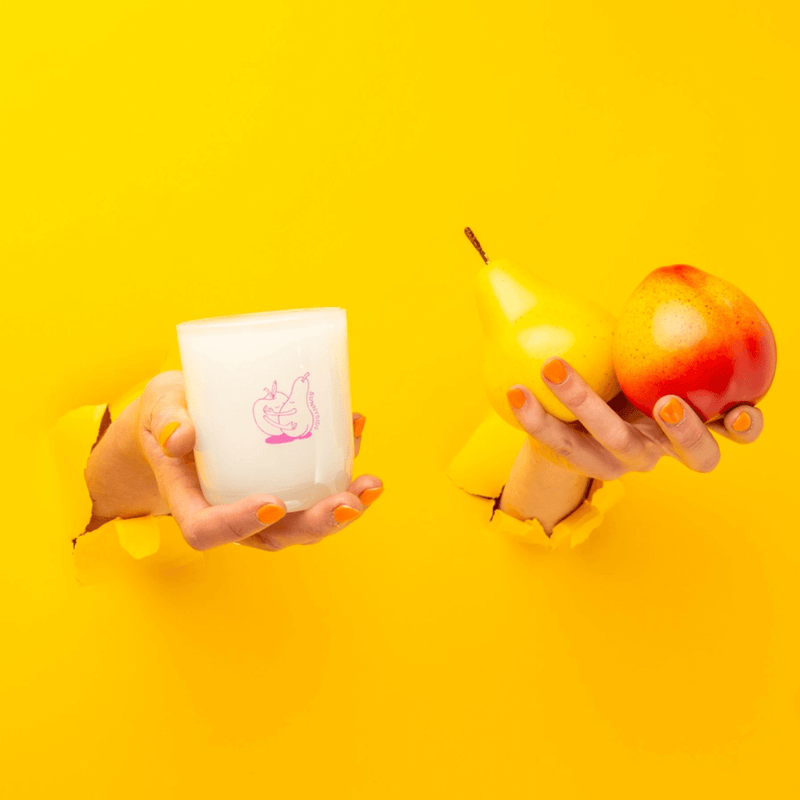 Hands holding Milkjar Sunnyside candle and a pear and nectarine through torn yellow paper.
