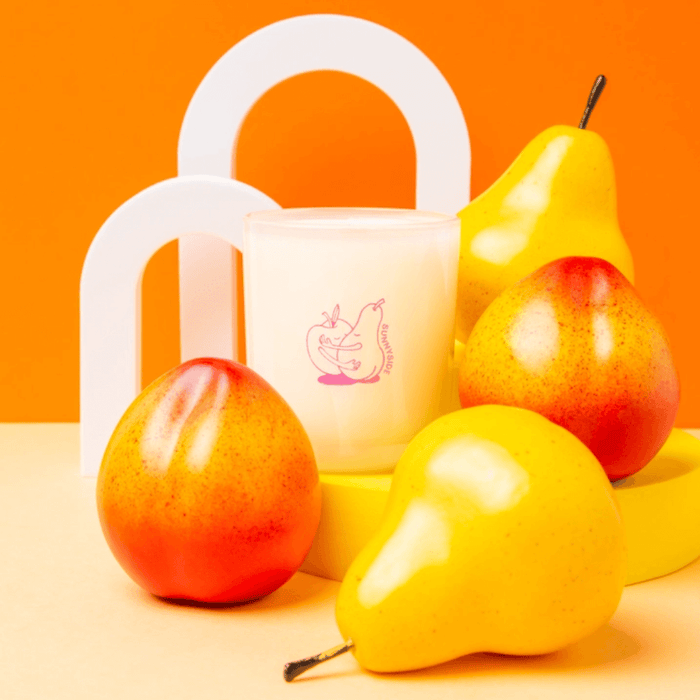 Styled photo of Milkjar Sunnyside candle with pears and nectarines on a yellow set with orange background.