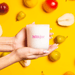 Hands holding the Milkjar Sunnyside candle against a yellow background with pears and stone fruit.