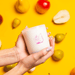 Hands holding the Milkjar Sunnyside candle with Milkjar branding against a yellow background with fruit.
