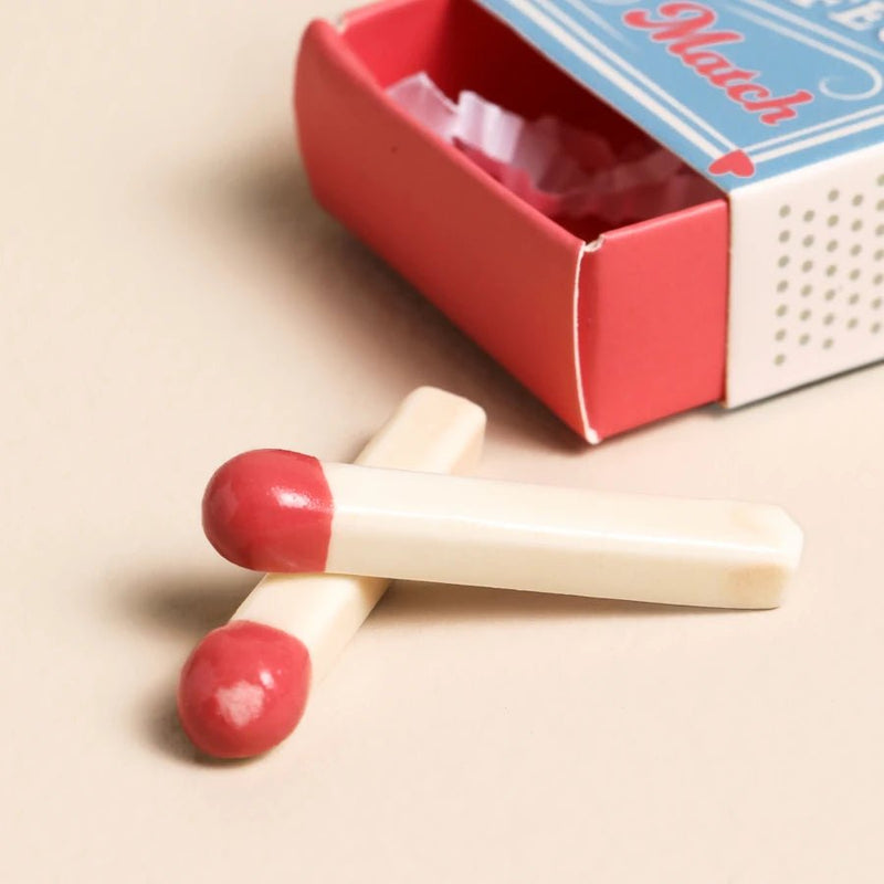 Two mini ceramic match tokens with red tips beside The Perfect Match matchbox