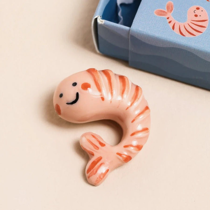 Pink glossy ceramic shrimp token beside the matching blue matchbox packaging