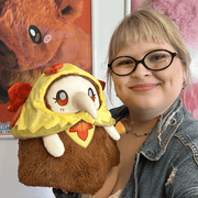 Person holding Mini Squishable Autumn Fairy Plague Nurse plush showing its fluffy brown body and autumn outfit
