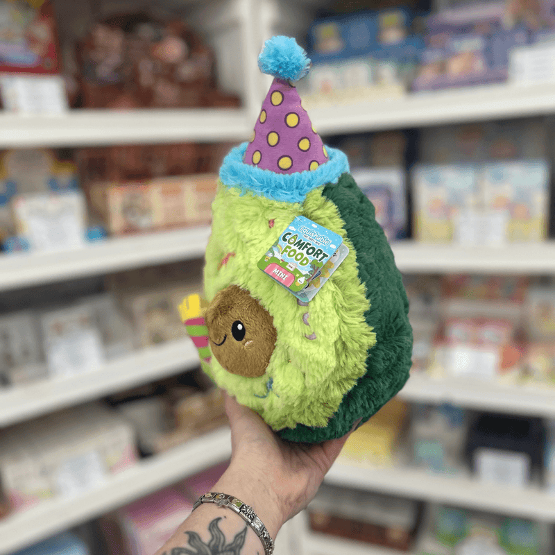 Mini Squishable Comfort Food Birthday Avocado plush toy held in hand, angled view with party hat and tag visible