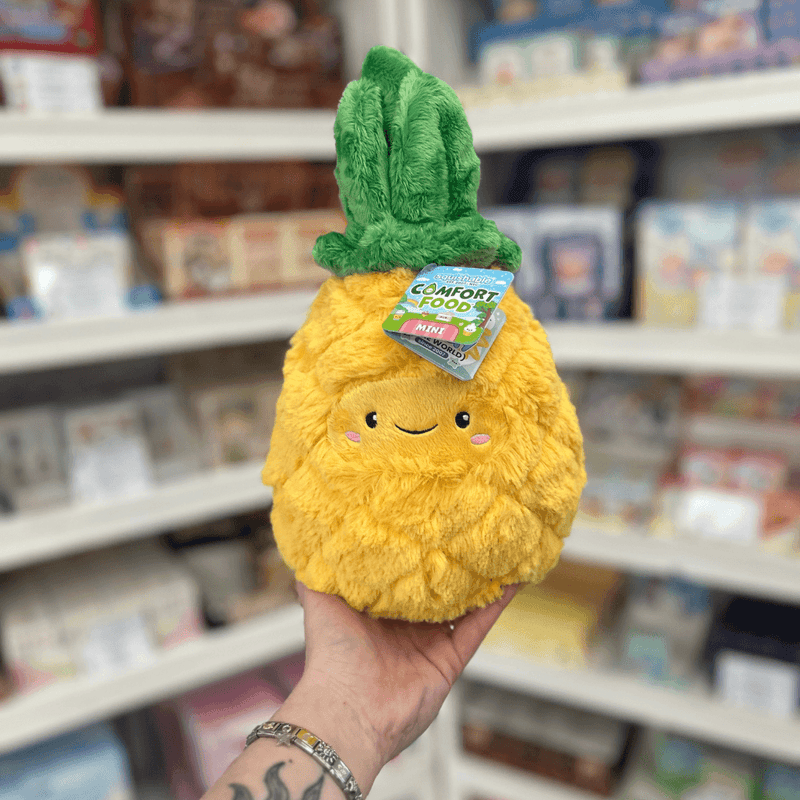 Mini Squishable Comfort Food Pineapple plush toy held in hand, front view showing the smiling pineapple face and soft green leaves